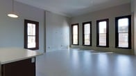 This single-floor space offers a clean, minimalist layout defined by its bright, open interior. The room is lined with multiple windows featuring dark wood frames, which allow generous amounts of natural light to filter in, illuminating the smooth, light-toned flooring throughout. The white walls provide a neutral backdrop, making the space highly adaptable for a range of uses. A structural column is positioned toward the center of the room, adding an architectural element without significantly interrupting the open floor plan. Crown molding runs along the ceiling, adding a subtle touch of traditional detail to the otherwise understated design. Along one side, a partial countertop or built-in surface is visible, hinting at potential utility or workspace functionality. The windows offer views of the surrounding urban streetscape, connecting the interior to its city environment. Overall, the space presents itself as a versatile, light-filled environment — essentially a blank canvas ready to be configured to suit a variety of residential or commercial purposes.