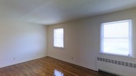 This single-floor space offers a clean, classic interior defined by its rich, polished hardwood floors that run throughout the main living area. The room is finished with smooth, neutral-toned walls that provide a bright and versatile backdrop, complemented by a white ceiling that enhances the sense of openness. Two windows allow generous amounts of natural light to filter into the space, creating a warm and inviting atmosphere throughout the day. A baseboard radiator unit is positioned beneath one of the windows, providing an efficient heat source during colder months. The layout is open and unobstructed, offering flexibility for a variety of furniture arrangements and uses. The overall aesthetic is simple and functional, with timeless architectural details that provide a solid foundation for personalization. The combination of gleaming hardwood floors, ample natural light, and a well-maintained interior makes this a practical and comfortable single-story space.