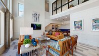 This dynamic two-story community amenity space is defined by its dramatic double-height ceilings and an open, airy architectural design that immediately commands attention. The main lounge area serves as the centerpiece, featuring soaring walls clad in shiplap-style paneling, a wall-mounted flat-screen TV, and an expansive arrangement of cognac leather sofas and colorful accent chairs set atop vibrant patterned rugs. Floor-to-ceiling windows flood the space with abundant natural light while offering direct views of the outdoor pool area, creating a seamless indoor-outdoor connection. An open staircase leads to a glass-railed upper mezzanine level, visible from below and adding architectural depth to the space. Adjacent to the main lounge, a dedicated game room features a full-size pool table, a wall-mounted TV, recessed and pendant lighting, and a built-in blue bench seat with bar-height seating — ideal for social gatherings. A separate co-working and lounge area is enclosed by large steel-framed glass partitions, offering a quieter retreat equipped with a work table, task seating, a flat-screen display, and comfortable sectional seating. Throughout, the design palette blends bold, eclectic artwork with contemporary furnishings, crown molding, and wood-look tile flooring to create an energetic yet welcoming communal environment.