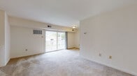 This single-floor residential unit offers a well-organized layout with clearly defined living, kitchen, sleeping, and bathroom areas. The living room serves as the central gathering space, finished with neutral wall-to-wall carpeting and crisp white walls that lend a clean, airy feel. A sliding glass door opens to an outdoor balcony, allowing natural light to flow in and providing views of the surrounding exterior. A staircase with a white half-wall railing is also visible within the living area, adding a subtle architectural detail. The kitchen is positioned adjacent to the living space and features warm wood-toned cabinetry, dark granite-style countertops, tile flooring, and black appliances including a full-size refrigerator and dishwasher. A stainless steel sink and ample upper and lower cabinet storage round out the functional cooking area. The bedrooms are carpeted and offer generous wall space, with sliding and hinged closet doors providing practical storage. Windows dressed with blinds allow natural light into the sleeping quarters. The bathroom is finished with tile flooring, a wood-toned vanity cabinet, stone-finish countertop, a white basin sink, and a well-lit mirror arrangement, creating a tidy and functional space.