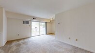 This single-floor residential unit offers a well-organized layout with clearly defined living, kitchen, sleeping, and bathroom areas. The living room serves as the central gathering space, finished with neutral wall-to-wall carpeting and crisp white walls that lend a clean, airy feel. A sliding glass door opens to an outdoor balcony, allowing natural light to flow in and providing views of the surrounding exterior. A staircase with a white half-wall railing is also visible within the living area, adding a subtle architectural detail. The kitchen is positioned adjacent to the living space and features warm wood-toned cabinetry, dark granite-style countertops, tile flooring, and black appliances including a full-size refrigerator and dishwasher. A stainless steel sink and ample upper and lower cabinet storage round out the functional cooking area. The bedrooms are carpeted and offer generous wall space, with sliding and hinged closet doors providing practical storage. Windows dressed with blinds allow natural light into the sleeping quarters. The bathroom is finished with tile flooring, a wood-toned vanity cabinet, stone-finish countertop, a white basin sink, and a well-lit mirror arrangement, creating a tidy and functional space.