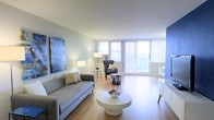 This well-appointed single-floor residence offers a thoughtfully designed open-concept living area that immediately impresses with its clean, modern aesthetic. The space is bathed in natural light, courtesy of large floor-to-ceiling windows dressed with vertical blinds, which also frame pleasant outdoor views and connect the interior to the surrounding environment.

The living room serves as the heart of the space, featuring warm hardwood flooring that runs throughout and lends a sense of continuity and warmth. A contemporary gray sofa anchors the seating area, complemented by an accent armchair and a stylish patterned ottoman. A round white coffee table serves as a central focal point, while a bold large-scale artwork adds a vibrant splash of color to the soft, neutral-toned walls.

Ambient and accent lighting, including coordinated floor and table lamps, ensure the space remains inviting at any time of day. The layout flows effortlessly, with a dining area visible toward the rear of the space, offering additional functionality. The overall ambiance is calm, airy, and sophisticated, striking a balance between practicality and refined style. Every element of the space has been curated to create a cohesive, comfortable, and visually appealing environment.