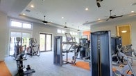 This single-floor fitness center offers a spacious, well-appointed workout environment designed to accommodate a variety of exercise needs. The facility is outfitted with professional-grade Matrix strength machines, cable systems, and cardiovascular equipment, including ellipticals and treadmills, strategically arranged to allow for comfortable movement throughout the space.

The room features a light grey color palette with a vibrant orange accent wall that adds an energizing touch to the overall ambiance. Recessed lighting throughout the ceiling is complemented by multiple ceiling fans, ensuring the space remains well-lit and well-ventilated during workouts. High transom windows line the upper walls, drawing in generous amounts of natural light and contributing to the open, airy feel of the room.

French doors on one end of the facility provide access to an outdoor area while further enhancing the flow of natural light into the space. The flooring combines durable, light-toned materials with orange equipment mats placed beneath key machines. The overall design strikes a balance between functionality and a clean, modern aesthetic.