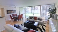 This single-floor space presents an open-plan layout that seamlessly integrates the living and dining areas into one cohesive environment. The living area is anchored by a pair of cream sofas accented with bold red and dark decorative cushions, arranged around a sleek black leather ottoman coffee table. A striking zebra-print area rug adds a dramatic visual contrast to the warm-toned parquet flooring that runs throughout the space.

The dining area sits adjacent to the living zone, featuring a wooden table surrounded by rich red-upholstered chairs, complemented by wall art and decorative accents that add character to the textured white brick wall. A ceiling fan overhead provides both practical airflow and a subtle design element.

The most commanding feature of the space is the expansive floor-to-ceiling sliding glass door system that spans nearly the entire rear wall, flooding the interior with abundant natural light and providing unobstructed views of a well-maintained green lawn outside. A decorative lattice panel to one side adds architectural interest. The overall ambiance is bold yet inviting, blending contemporary furnishings with warm natural tones.