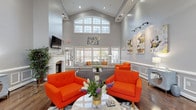 The Park Place at Woburn clubhouse is a thoughtfully designed community gathering space that masterfully blends classic architectural details with modern, energetic style. The main lounge commands attention with dramatic vaulted ceilings that soar overhead, anchored by a large arched window that floods the room with natural light. Elegant wainscoting lines the soft gray walls throughout, lending a refined, traditional character to the interior.

The lounge area is furnished with bold orange accent chairs and neutral gray sofas accented with vibrant throw pillows, all arranged around a herringbone-patterned area rug atop rich hardwood floors. A white-mantled fireplace serves as the room's focal point, with a wall-mounted television above it, creating an inviting atmosphere for relaxation and socializing. Contemporary artwork displayed on track-lit gallery walls adds a creative, curated touch.

A dedicated office area, framed by white decorative columns, provides a professional workspace complete with desks and a reception counter — ideal for leasing and administrative functions. The entry foyer features glass-paned double doors that open to outdoor views, enhancing the connection between interior and exterior. Together, these spaces create a welcoming, vibrant, and functional environment for residents and visitors alike.