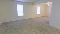 This single-floor space presents a straightforward and functional layout designed for comfortable living. The main living area is generously proportioned, featuring wall-to-wall carpeting in a neutral beige tone that adds warmth underfoot and contributes to the overall understated aesthetic of the interior. The walls are finished in a light cream color, creating a bright and airy atmosphere throughout. Two windows fitted with white horizontal blinds allow natural light to filter into the space, keeping the room well-lit while offering privacy and light control as needed. The ceiling is smooth and consistent in height, lending a clean, uncluttered feel to the room. A subtle architectural detail is visible in the upper corner of the space, suggesting a transition or partial partition between areas, which may define distinct zones within the open floor plan. The overall ambiance is calm and neutral, providing a versatile backdrop that can be easily personalized. The space is well-suited for a variety of uses and offers a solid foundation for anyone looking to make it their own.