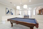 This well-appointed single-floor space combines functional design with refined aesthetics throughout its key areas. The kitchen is a standout feature, showcasing floor-to-ceiling white shaker cabinetry, a classic subway tile backsplash, and stainless steel appliances including a French door refrigerator, microwave, and range. A large central island with a crisp white countertop serves as both a prep area and a casual dining spot, complemented by stylish blue-and-white patterned counter-height chairs. Warm wood-look tile flooring and elegant pendant lighting add to the inviting atmosphere, while natural light filters in through windows dressed with decorative curtains and rich mahogany-toned doors provide a warm contrast to the bright, neutral palette.

The expansive game room is equally impressive, featuring herringbone-patterned tile flooring, crown molding, and recessed lighting throughout. A full-size pool table anchors the space beneath a cluster of drum pendant lights, while a cozy round seating arrangement with upholstered blue chairs provides a comfortable gathering area. A wall-mounted flat-screen television, arched architectural niches, and a pair of bold abstract artworks contribute to the room's sophisticated character. The open layout and seamless flow between areas make this space ideal for both entertaining and everyday living.