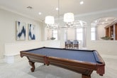 This well-appointed single-floor space combines functional design with refined aesthetics throughout its key areas. The kitchen is a standout feature, showcasing floor-to-ceiling white shaker cabinetry, a classic subway tile backsplash, and stainless steel appliances including a French door refrigerator, microwave, and range. A large central island with a crisp white countertop serves as both a prep area and a casual dining spot, complemented by stylish blue-and-white patterned counter-height chairs. Warm wood-look tile flooring and elegant pendant lighting add to the inviting atmosphere, while natural light filters in through windows dressed with decorative curtains and rich mahogany-toned doors provide a warm contrast to the bright, neutral palette.

The expansive game room is equally impressive, featuring herringbone-patterned tile flooring, crown molding, and recessed lighting throughout. A full-size pool table anchors the space beneath a cluster of drum pendant lights, while a cozy round seating arrangement with upholstered blue chairs provides a comfortable gathering area. A wall-mounted flat-screen television, arched architectural niches, and a pair of bold abstract artworks contribute to the room's sophisticated character. The open layout and seamless flow between areas make this space ideal for both entertaining and everyday living.