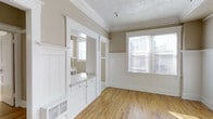 This well-appointed 1-bedroom space showcases a blend of classic craftsmanship and timeless design. The interior is defined by elegant crown molding, crisp white wainscoting that lines the lower walls, and warm-toned hardwood floors that extend throughout, lending the space a welcoming and cohesive feel.

Natural light flows generously through the windows, fitted with blinds for privacy and light control, illuminating the neutral taupe wall tones that create a calm, understated backdrop. The textured ceiling adds a traditional character that complements the careful millwork details found throughout.

A built-in cabinetry nook with a countertop and small drawers provides practical storage and workspace, seamlessly integrated into the architecture with white painted finishes that maintain visual continuity. Decorative white columns and framed openings define the transition between spaces, adding a sense of structure and formality while keeping the layout open and connected.

A floor-level vent indicates a central heating system integrated discreetly into the design. Overall, the space exudes a classic residential charm, combining period-appropriate architectural details with a clean, neutral palette that offers a versatile and inviting atmosphere.