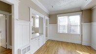 This well-appointed 1-bedroom space showcases a blend of classic craftsmanship and timeless design. The interior is defined by elegant crown molding, crisp white wainscoting that lines the lower walls, and warm-toned hardwood floors that extend throughout, lending the space a welcoming and cohesive feel.

Natural light flows generously through the windows, fitted with blinds for privacy and light control, illuminating the neutral taupe wall tones that create a calm, understated backdrop. The textured ceiling adds a traditional character that complements the careful millwork details found throughout.

A built-in cabinetry nook with a countertop and small drawers provides practical storage and workspace, seamlessly integrated into the architecture with white painted finishes that maintain visual continuity. Decorative white columns and framed openings define the transition between spaces, adding a sense of structure and formality while keeping the layout open and connected.

A floor-level vent indicates a central heating system integrated discreetly into the design. Overall, the space exudes a classic residential charm, combining period-appropriate architectural details with a clean, neutral palette that offers a versatile and inviting atmosphere.