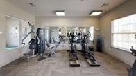 This fitness center is designed to support a well-rounded workout routine, offering a thoughtful mix of cardio and strength training equipment within a clean, single-floor layout. The space features neutral-toned walls, patterned carpet tile flooring, and flush-mounted ceiling lighting that keeps the environment bright and functional. Large mirrors line key wall sections, visually expanding the room while allowing users to monitor their form during exercises.

The cardio area is equipped with treadmills, elliptical trainers, and a stationary bike, alongside a standing scale for tracking progress. Natural light filters in through multiple windows fitted with horizontal blinds, complementing the overhead lighting. A wall-mounted flat-screen television provides entertainment during workouts.

The strength training section includes a multi-station cable machine with a full exercise chart, a dedicated free weight rack stocked with a range of dumbbells, and additional weight benches. A water cooler is conveniently positioned nearby. A glass-paneled door connects the fitness center to an adjacent room, adding to the open and accessible feel of the overall space.