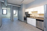 This single-floor unit showcases a compelling blend of industrial and modern design. Polished concrete floors run throughout the space, complementing the exposed concrete ceilings and structural columns that reflect its loft-style character. The open living area benefits from an airy, expansive feel, with visible ductwork and ceiling infrastructure adding to the industrial aesthetic. Arched architectural details and a prominent cylindrical column provide visual interest and character.

The kitchen features white shaker-style cabinetry with brushed hardware, dark countertops, and a stainless steel appliance suite including a refrigerator, dishwasher, electric range, and over-range microwave. A double-basin sink anchors the layout, which offers generous counter and storage space.

The bedroom includes a walk-in closet, while an additional room with exposed white brick walls and dark-framed windows brings natural light and texture to the space. The bathroom is finished with large-format tile flooring, a walk-in shower with a curtain rod, and a white vanity. Throughout, gray-painted walls and white trim create a cohesive, neutral palette suited to a variety of uses.