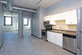 This single-floor unit showcases a compelling blend of industrial and modern design. Polished concrete floors run throughout the space, complementing the exposed concrete ceilings and structural columns that reflect its loft-style character. The open living area benefits from an airy, expansive feel, with visible ductwork and ceiling infrastructure adding to the industrial aesthetic. Arched architectural details and a prominent cylindrical column provide visual interest and character.

The kitchen features white shaker-style cabinetry with brushed hardware, dark countertops, and a stainless steel appliance suite including a refrigerator, dishwasher, electric range, and over-range microwave. A double-basin sink anchors the layout, which offers generous counter and storage space.

The bedroom includes a walk-in closet, while an additional room with exposed white brick walls and dark-framed windows brings natural light and texture to the space. The bathroom is finished with large-format tile flooring, a walk-in shower with a curtain rod, and a white vanity. Throughout, gray-painted walls and white trim create a cohesive, neutral palette suited to a variety of uses.