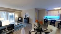 This single-floor residence offers a thoughtfully designed open-concept layout that effortlessly connects the living, dining, and kitchen areas into one cohesive, functional space. The interior is finished with smooth light gray walls and durable wood-look flooring that runs throughout, lending a clean, contemporary aesthetic to the entire space.

The dining area is positioned at the heart of the layout, centered around a round dining table set with elegant place settings, glassware, and a vibrant floral centerpiece that adds a warm, inviting touch. White upholstered dining chairs complement the refined yet approachable atmosphere.

Flowing naturally from the dining space, the living area is furnished with a plush accent chair in deep navy blue, accented by a patterned area rug in coordinating tones. A stylish black dresser with decorative accents serves as a focal point, while a floor lamp provides warm ambient lighting. Large windows along the far wall allow natural light to fill the space.

The kitchen, partially visible from the main living area, features a breakfast bar with seating and illuminated cabinetry, hinting at a modern and well-equipped culinary space. A hallway leads to additional private areas of the home, completing this well-balanced and move-in-ready residence.