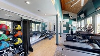 Located at 575 Matmor Rd in Woodland, CA, this fitness center is a thoughtfully designed exercise facility that combines functionality with a modern aesthetic. The space features soaring vaulted ceilings accented with warm wood paneling and bold teal walls adorned with geometric patterns, creating an energizing atmosphere for workouts.

The exercise room is comprehensively equipped with a row of treadmills, elliptical trainers, a stationary spin bike, a rowing machine, and a cable strength machine, complemented by a full dumbbell rack with a range of free weights. Structural columns integrated into the layout provide visual separation between zones without interrupting the open flow of the space.

Flooring transitions between light hardwood and patterned carpet tiles, delineating cardio and strength training areas. Large windows along one wall flood the room with natural light and offer direct views of an outdoor pool area, enhancing the sense of openness. Full-length mirrors along the opposite wall visually expand the space and support proper form during workouts.

Practical amenities include a wall-mounted water fountain with a sanitation station, ceiling fans for air circulation, mounted flat-screen televisions, and recessed lighting. Decorative touches such as woven circular wall art and indoor plants add warmth, making this facility both functional and inviting.