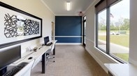 This single-floor office space offers a clean, contemporary design that balances functionality with a polished aesthetic. The layout accommodates multiple workstations arranged along sleek white desks with modern ergonomic chairs, making it well-suited for a collaborative or shared work environment. A striking deep navy blue accent wall serves as the focal point of the space, accented by a crisp white chair rail that adds a refined architectural detail. The remaining walls are finished in a neutral off-white tone, creating a bright and professional atmosphere. Large windows with dark-framed roller blinds line the perimeter, flooding the space with natural light and offering open views of a lush green landscape beyond. The flooring throughout consists of durable patterned carpet tiles in warm neutral tones, adding texture and comfort underfoot. Overhead, linear fluorescent fixtures provide consistent, even illumination. Tasteful wall art — including a bold black-and-white abstract piece — adds a creative touch to the workspace. A glass-paneled door connects to an adjacent area, enhancing the sense of openness and flow within the overall floor plan. Crown molding along the ceiling line contributes a subtle but elegant finishing detail throughout the space.