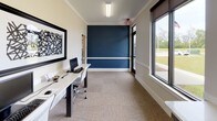 This single-floor office space offers a clean, contemporary design that balances functionality with a polished aesthetic. The layout accommodates multiple workstations arranged along sleek white desks with modern ergonomic chairs, making it well-suited for a collaborative or shared work environment. A striking deep navy blue accent wall serves as the focal point of the space, accented by a crisp white chair rail that adds a refined architectural detail. The remaining walls are finished in a neutral off-white tone, creating a bright and professional atmosphere. Large windows with dark-framed roller blinds line the perimeter, flooding the space with natural light and offering open views of a lush green landscape beyond. The flooring throughout consists of durable patterned carpet tiles in warm neutral tones, adding texture and comfort underfoot. Overhead, linear fluorescent fixtures provide consistent, even illumination. Tasteful wall art — including a bold black-and-white abstract piece — adds a creative touch to the workspace. A glass-paneled door connects to an adjacent area, enhancing the sense of openness and flow within the overall floor plan. Crown molding along the ceiling line contributes a subtle but elegant finishing detail throughout the space.