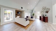 This well-appointed single-floor home showcases an open-concept layout designed for both comfortable living and effortless entertaining. The expansive living room immediately draws attention with its dramatic vaulted ceilings and a striking floor-to-ceiling stacked stone fireplace, complete with a wood mantel and wall-mounted television. Soft sage-green walls, light hardwood flooring, and recessed lighting create a warm, inviting ambiance throughout. Large windows and French doors flood the space with natural light and provide views of the surrounding greenery, while also offering seamless access to the outdoors.

The dining area is a standout feature, anchored by a solid wood dining table and cross-back chairs set atop a natural jute rug. A generous bank of windows frames lush garden views, making every meal an immersive experience.

The kitchen is thoughtfully designed with crisp white cabinetry, granite countertops, stainless steel appliances including a side-by-side refrigerator, and a distinctive blue mosaic tile backsplash that adds a touch of visual interest. A dedicated bar seating area connects to the main living spaces, enhancing the home's open flow.

Throughout, the consistent use of light tones, natural materials, and considered architectural details — including high ceilings carried across multiple rooms — give this home a spacious, airy character.