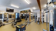 This fitness center combines rustic charm with modern functionality, creating an inviting and practical space for exercise. The interior is defined by prominent exposed wooden beams and framing elements that add warmth and character to the otherwise clean, white walls and ceilings. Bright fluorescent lighting ensures the entire space is well-illuminated throughout the day.

The gym floor is finished with durable, cushioned flooring suited for high-impact activity, and the layout is thoughtfully organized to accommodate a diverse range of workout routines. A dedicated free weights area features a full rack of dumbbells in varying sizes, accompanied by adjustable benches for seated and lying exercises. Plyo boxes are also present, catering to functional and high-intensity training.

The strength training section includes a multi-station cable and pulley machine, ideal for a wide variety of resistance exercises. Cardio equipment, including treadmills and other aerobic machines, lines the perimeter of the space. Wall-mounted mirrors expand the sense of space and allow users to monitor their form during workouts. A mounted flat-screen television adds an entertainment element to the cardio area. The overall ambiance is energetic yet welcoming, blending natural materials with modern fitness amenities.