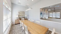 Located at 4832 Ruby Red Drive in Chattanooga, TN, this conference room offers a polished and welcoming environment well-suited for meetings and collaborative gatherings. The room is defined by its soaring ceilings and soft, neutral color palette of light gray and white, creating an open and airy atmosphere throughout.

At the center of the space sits a substantial natural wood conference table surrounded by white slatted chairs, comfortably accommodating up to eight attendees. A sleek linear pendant light hangs directly above the table, providing focused, modern illumination complemented by recessed ceiling lights. A textured area rug anchors the seating area, adding warmth and visual interest to the flooring beneath.

Natural light filters into the room through a set of windows fitted with white horizontal blinds, while a pass-through window connects the conference room to an adjacent amenity area visible in the background. French-style doors with glass panels provide an elegant entry point and allow borrowed light from neighboring spaces.

A decorative sideboard with a geometric wood-inlay pattern lines one wall, topped with matching table lamps and a large abstract landscape artwork, lending a sophisticated, residential quality to the overall design. The combination of functional furnishings and refined décor makes this conference room a comfortable and professional setting.