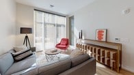 This single-floor residence showcases a thoughtfully designed, modern interior with a cohesive contemporary aesthetic throughout. The open-concept living area is bathed in natural light from large floor-to-ceiling windows fitted with roller shades, creating an airy atmosphere. The space accommodates a sectional sofa, accent seating, and a sculptural wood-and-metal shelving unit, while warm hardwood flooring runs throughout the main living areas.

The kitchen is outfitted with sleek, dark matte cabinetry extending to the ceiling, complemented by white quartz countertops and a white subway tile backsplash. A peninsula with bar seating serves as both a workspace and casual dining area. Stainless steel appliances, including a refrigerator, range, microwave, and dishwasher, complete the functional layout.

The bedroom features soft carpet underfoot, angled ceilings that add architectural interest, and access to both a dedicated closet with built-in shelving and an adjacent bathroom. The bathroom is finished with hexagonal white floor tile, a floating wood-toned vanity with a white countertop, and a tub-shower combination. Throughout the unit, neutral tones and clean lines create a calm, well-balanced living environment.