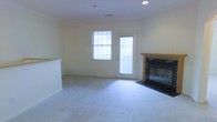 This two-story space presents a well-organized layout designed for comfortable everyday living. The main living area, visible in the image, is finished with plush wall-to-wall carpeting that adds warmth underfoot. Neutral-toned walls and recessed lighting contribute to a clean, understated aesthetic throughout. A wood-mantled fireplace with a dark surround serves as a natural focal point in the living room, providing both a functional heating element and a classic architectural detail. Natural light enters generously through a multi-pane window alongside a glass-paneled exterior door, brightening the space and offering direct outdoor access. Crown molding frames the ceiling, adding a subtle traditional touch to the overall design. The open feel of the living area is enhanced by its generous proportions, with the partial view of a staircase railing indicating the connection to the second floor. The second level expands the overall footprint of the space, providing additional room separation and privacy. The combination of thoughtful architectural detailing, natural light, and practical features like the fireplace and outdoor access makes this a well-rounded and inviting space.