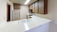 This single-floor unit in La Mesa offers a straightforward, functional layout with clearly defined living spaces. The living room is generously proportioned, featuring neutral-toned carpet, a textured ceiling, and a wall-mounted air conditioning unit. Vertical blinds cover a sliding glass door that allows natural light to fill the room. Adjacent to the living area, the dining space is distinguished by a chandelier overhead and direct access to the kitchen via a pass-through breakfast bar with a light-colored countertop.

The kitchen is equipped with dark wood cabinetry providing ample storage, a refrigerator, electric range, built-in microwave, and a double-basin sink set into a speckled white countertop. The layout is efficient and functional. The bedrooms are carpeted with neutral walls, textured ceilings, and windows fitted with vertical blinds that bring in outdoor greenery views. The bathroom features wood-look flooring, a wood-tone vanity with a stone-patterned countertop, a full tub-shower combination, and clean white fixtures. White baseboards run consistently throughout, contributing to a cohesive and tidy overall appearance.