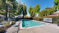 Located at 455 Brahms Way in Sunnyvale, California, this outdoor pool area serves as a central community amenity designed for relaxation and recreation. The generously sized swimming pool features clear, blue-green water and is finished with classic tiled edges, offering an inviting atmosphere under the California sun. The surrounding pool deck provides ample space for lounging, with chaise lounge chairs and patio seating conveniently arranged for residents to enjoy. Shaded umbrella tables are positioned nearby, offering a comfortable retreat from the sun during warm days. The area is enclosed by a wrought iron fence with integrated greenery and manicured shrubs, creating a sense of privacy while maintaining an open, airy feel. Towering mature trees, including striking redwoods, frame the space and cast dappled shade across the deck, enhancing the natural ambiance. The lush, well-kept landscaping adds vibrant greenery that complements the sunny, blue-sky backdrop characteristic of the South Bay. Residential buildings with neutral-toned, multi-pitched rooflines are visible in the background, providing a sense of the broader community context. This outdoor space combines functional amenities with a tranquil, park-like setting, making it a standout feature of the property.