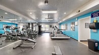 This fitness center is a comprehensively outfitted workout facility spread across a single floor, offering ample space for a wide variety of exercise routines. The main workout area is defined by its vibrant teal accent walls, which contrast with the neutral white surfaces and textured ceilings to create an energetic yet focused atmosphere. Ceiling fans and recessed lighting, along with fluorescent light fixtures, ensure the space is well-lit throughout.

The cardio section features a row of treadmills and additional cardio machines, including stationary bikes and ellipticals, positioned along one side of the room for easy access. On the opposite side, a full wall of mirrors visually expands the space and allows users to monitor their form during workouts. The strength-training area includes a multi-station cable machine and a variety of resistance equipment, while a well-organized free weight rack holds an assortment of dumbbells for functional training.

Durable rubber flooring covers the entire workout area, providing both safety and comfort. Natural light filters in through windows, offering views to the outdoors and enhancing the overall ambiance. An adjoining room provides additional space, likely for storage or administrative use. The fitness center is thoughtfully arranged to maximize functionality while maintaining an open, accessible layout.