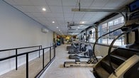 This fitness facility is designed to support a comprehensive workout experience within a long, efficiently organized single-floor space. The layout features a row of cardio machines — including treadmills, ellipticals, and a WaterRower — positioned along one side, allowing users ample room to exercise comfortably. A dedicated area for functional training is visible toward the rear, stocked with medicine balls and other accessories mounted on a wall-mounted rack, adding versatility to workout options. The dropped ceiling with recessed lighting provides consistent, even illumination throughout the space, while a wall-mounted split air conditioning unit ensures climate control. Natural light filters in through a large garage-style window on one end, offering a view of the surrounding exterior. Light-toned flooring with a subtle texture runs the length of the room, complementing the neutral gray walls and creating a clean, focused atmosphere. A railing system along one side adds a structural design element while contributing to safe navigation through the space. Overall, the facility presents a practical and well-maintained environment suited for a variety of fitness routines.