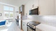 Located at 1457 N Main St in Los Angeles, CA, this efficiently designed single-floor unit offers a well-organized layout comprising a living area, kitchen, and full bathroom. The living space is bright and airy, highlighted by large floor-to-ceiling windows that flood the room with natural light and offer open views of the surrounding neighborhood. Light wood-tone flooring runs throughout, complementing the clean, white walls and creating a cohesive, modern aesthetic. The living area provides ample room for a bed, storage cabinetry, and a small dining setup, making smart use of the available space. The kitchen is thoughtfully appointed with two-tone cabinetry — white uppers and light grey lowers — paired with a white subway tile backsplash and light quartz countertops. Stainless steel appliances include a range, over-the-range microwave, dishwasher, and refrigerator, offering full functionality in a compact footprint. The bathroom features a minimalist design with light wood flooring, a white vanity with quartz countertop, and a backlit LED mirror that adds a modern, polished touch. The overall ambiance of the space is clean, contemporary, and practical, making it a well-suited unit for urban living in Los Angeles.