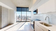Located at 81 E 14th St in Kansas City, MO, this single-floor unit is situated on an upper level of a modern high-rise building, offering commanding panoramic views of the Kansas City skyline. The architecture emphasizes a clean, contemporary aesthetic throughout, with exposed concrete ceilings, light hardwood-style flooring, and an open, airy layout that maximizes both space and natural light.

The main living area is anchored by dramatic floor-to-ceiling windows that frame sweeping city views and flood the interior with abundant sunlight. The open-concept kitchen is sleekly designed, featuring a combination of white upper cabinetry and warm-toned lower cabinets, a marble-look tile backsplash, white quartz countertops, and a full suite of stainless steel appliances including a range, microwave, dishwasher, and refrigerator. The kitchen flows seamlessly into the living space, reinforcing the unit's cohesive, modern character.

The bathroom is finished with large-format marble-look floor tiles, a glass-enclosed shower, a vessel sink atop a wood-framed vanity with white cabinetry, and a large mirror. A stacked in-unit washer is conveniently integrated into the bathroom space with dedicated cabinetry above. Light hardwood-style flooring runs consistently throughout the living areas, contributing to the unit's bright, contemporary ambiance.