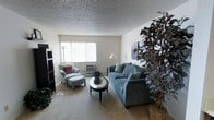 This single-floor space centers around a well-appointed living room designed for comfortable everyday use. The room is finished with wall-to-wall carpeting in a neutral tone, contributing to a warm and inviting atmosphere. A textured popcorn ceiling adds a classic architectural detail overhead.

A large window fitted with vertical blinds serves as the focal point of one wall, drawing in an abundance of natural light and brightening the interior. Beneath the window, a wall-mounted air conditioning unit provides climate control directly within the living area.

The living room is furnished with a full-sized sofa in a muted blue-grey upholstery, paired with a coordinating armchair and ottoman set accented by a red throw pillow. A round mirrored coffee table anchors the seating arrangement, adding a reflective decorative element. A tall indoor plant positioned near the seating area introduces a natural, organic touch to the space.

Additional details include a tall floor lamp and a table lamp providing supplemental lighting, a wall-mounted art piece, and a media storage unit along one wall. The overall ambiance is calm and functional, with a traditional residential character.