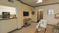 This single-story space showcases a thoughtfully designed interior with a cohesive neutral color palette of cream and warm beige tones throughout. The living area serves as the central gathering space, featuring plush carpeted flooring, bold dark baseboards and crown molding that frame the room with a polished, finished look. A wall-mounted flat-screen television is positioned above a slim wooden console table, creating a clean and functional focal point for the room.

A ceiling fan with integrated lighting provides both comfort and illumination to the open space. An arched doorway adds an elegant architectural detail, leading into a hallway with a dark-painted door, lending character to the layout. The living area connects seamlessly to the kitchen, which is visible through an open pass-through or partial wall, featuring white cabinetry and a raised bar-style counter. The kitchen's proximity to the living area enhances the open, interconnected flow of the space.

The overall ambiance is warm and inviting, with understated coastal-inspired décor accents, including decorative pillows, candle holders, and natural textures, contributing to a comfortable, casual atmosphere.