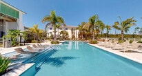 Located at 6026 Whitaker Rd in Naples, Florida, this expansive outdoor amenity space embodies the essence of Florida resort living. At its centerpiece sits a generously sized swimming pool with crystal-clear blue water, complemented by in-pool loungers and entry steps that invite leisurely relaxation. A shallow sun shelf provides an ideal spot for lounging partially submerged, while the surrounding paver deck is lined with contemporary chaise loungers designed for sunbathing in the abundant Florida sunshine.

The landscaping is a standout feature, with tall swaying palm trees, tropical foliage, agave plants, and manicured garden beds creating a lush, resort-like atmosphere throughout the space. Turfed lawn areas provide additional lounging zones, shaded by large umbrellas for those seeking a respite from the sun. Decorative tiled planters and modern amenity fixtures add refined detail to the hardscape.

Framing the pool area are multi-story white residential buildings with balconies and green architectural accents, while the perimeter opens to tranquil waterfront and natural tree line views beyond the fencing. A clubhouse-style structure with large windows and a covered veranda anchors one side of the space, enhancing the social and recreational character of this vibrant outdoor retreat.