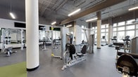 This fitness center offers a well-equipped, open-plan workout space designed to accommodate a wide range of exercise needs. The facility features soaring ceilings with exposed ductwork and industrial-style linear fluorescent lighting, lending the space a contemporary, loft-like aesthetic. Large windows along the perimeter walls allow abundant natural light to fill the interior, creating a bright and energizing environment.

The flooring throughout is a durable rubber material in a neutral gray tone, providing both safety and comfort during workouts. Sturdy cylindrical columns are positioned throughout the space, complementing the architectural character of the facility. Full-length mirrors are strategically placed to allow users to monitor their form during exercise.

The equipment selection is comprehensive, including Hoist-brand strength machines, a cable system, and cardio equipment such as treadmills. A dedicated free weights area with a full dumbbell rack is visible along one wall. The layout is thoughtfully organized to ensure ample circulation space between stations, supporting a safe and efficient workout experience for multiple users simultaneously.
