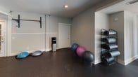 This fitness center is thoughtfully designed to support a comprehensive workout experience across two connected zones. The main cardio area is bright and airy, benefiting from large windows that frame views of an outdoor pool, filling the space with natural light and creating an open, energizing atmosphere. Recessed ceiling lighting supplements throughout, ensuring consistent illumination. The rubber flooring throughout the entire facility provides both durability and comfort underfoot.

The cardio zone is outfitted with a lineup of professional-grade machines, including treadmills, a StairMaster, and ellipticals, positioned alongside floor-to-ceiling mirrors that visually expand the space. A dedicated weight training area features a tiered dumbbell rack with a full range of free weights, multiple adjustable benches, and additional strength equipment, all organized for efficient use.

An adjoining room offers a versatile functional training space with rubber flooring, wall-mounted pull-up bars, stability and exercise balls, a yoga mat storage rack, and a water dispenser—ideal for stretching, yoga, or dynamic workouts. The overall aesthetic is clean and neutral, with light gray walls, white trim, and a well-maintained, professional environment.