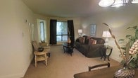 This single-floor residence offers a comfortable and well-appointed living space designed with a warm, neutral color palette throughout. The living room serves as the heart of the home, featuring soft wall-to-wall carpeting that adds a cozy, inviting atmosphere underfoot. Large windows dressed with floor-length dark drapes allow for flexible light control while framing views of the surrounding greenery, filling the room with natural light when open. The space is thoughtfully arranged with generous seating, including a plush dark sofa accented with warm-toned throw pillows, a classic round coffee table as a central focal point, and additional accent seating that adds character to the layout. Ambient lighting is provided by elegant table lamps positioned throughout the room, creating a soft and welcoming glow. A framed piece of wall art adds a decorative touch, while the smooth, neutral-toned walls provide a versatile backdrop for any style of furnishing. An adjacent area is visible through a doorway, suggesting a well-connected floor plan that flows naturally between spaces. Overall, the residence presents a clean, livable design with practical features and a comfortable ambiance suited for everyday living.
