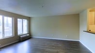 This single-floor space features an open living area designed with a clean, functional layout. The room is finished with dark hardwood-style flooring that contrasts neatly against the warm beige walls and crisp white baseboards, creating a simple yet welcoming aesthetic. A large window along one wall allows natural light to fill the space, enhancing its open feel. A wall-mounted PTAC heating and cooling unit is positioned beneath the window, providing climate control directly within the room. The ceiling is smooth and features a sprinkler head, indicating a modern fire safety system. Multiple electrical outlets are evenly distributed along the walls, offering practical convenience throughout the space. The neutral color palette and unfurnished layout provide a versatile canvas suitable for a variety of arrangements and uses.