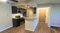 This single-floor unit is designed with modern finishes and functional layouts throughout. The kitchen serves as a central highlight, featuring dark espresso cabinetry paired with granite countertops, a tile backsplash, and a full suite of stainless steel appliances including a range, microwave, and refrigerator. A peninsula with an integrated sink and matching granite surface provides additional prep space and casual seating, while track lighting overhead ensures the area is well-lit. Hardwood-style flooring runs throughout, adding warmth and continuity to the living spaces. The bathroom is equally well-appointed, showcasing a wide double-sink vanity topped with a granite countertop and fitted with brushed nickel faucets and towel ring hardware. Dark cabinetry beneath the dual sinks provides ample storage, while large framed mirrors above each sink enhance the sense of space. Neutral wall tones throughout the unit create a clean, versatile backdrop that complements the rich, dark cabinetry and metallic fixtures. The overall aesthetic balances contemporary design with practical functionality, making this a comfortable and stylish living space.