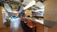 Located in Chicago's South Loop neighborhood, this single-floor space at 343 S Dearborn St offers a distinctive blend of industrial character and eclectic interior design. Entry is made through a set of sleek, glass-paneled double doors framed in aluminum, opening onto hardwood-floored corridors and a polished concrete main floor.

The space is defined by its exposed structural beams painted in deep green, visible ductwork, and a mix of concrete and hardwood flooring that gives it an industrial yet inviting atmosphere. A central games area features a bold yellow billiards table beneath pendant lighting, while bold floral wallpaper adds a dramatic visual contrast to the raw ceiling elements.

A well-appointed bar counter with a dark granite top and upholstered bar stools serves as a central social hub, adjacent to a fully equipped kitchenette outfitted with oak cabinetry, dark countertops, a sink, microwave, and dishwasher. A skylight introduces natural light into the space.

Multiple lounge seating areas are thoughtfully arranged throughout, including a cozy TV lounge with a slate tile accent wall, exposed beam ceiling, a round stone coffee table, and plush seating. Another seating nook features a rich blue velvet sofa set atop a teal area rug. The space is well-suited for private events, gatherings, or entertainment.