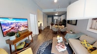 This single-floor residence showcases an open-concept floor plan that seamlessly connects the living, dining, and kitchen areas into one cohesive living space. The hardwood-style flooring runs throughout, lending warmth and continuity across each zone.

The living area is comfortably sized, anchored by a plush sofa, a patterned area rug, and a wooden coffee table. A media console supports a large flat-screen television, completing the entertainment setup. Track lighting overhead provides adjustable illumination throughout the main space.

The dining area sits centrally within the open layout, featuring a round table surrounded by cushioned accent chairs — an inviting arrangement suited for casual dining or entertaining.

The kitchen is positioned at the rear and features white cabinetry, a dark countertop island or breakfast bar, and stainless steel appliances including a full-sized refrigerator. The design is clean and functional, with ample cabinet storage.

A hallway visible near the entry suggests access to additional private rooms. The overall ambiance is modern and welcoming, with a neutral color palette accented by blue-toned walls in the kitchen area, creating a stylish, well-organized living environment.