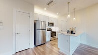 This well-appointed single-floor unit offers a thoughtfully designed open-concept layout that seamlessly connects the kitchen and living areas. The kitchen is a standout feature, showcasing crisp white shaker-style cabinetry extending to the ceiling, a full suite of stainless steel appliances including a refrigerator, electric range, and microwave, and a central island with a light quartz countertop and integrated sink. Elegant glass pendant lights suspended from above add a refined touch to the space. Light wood-look plank flooring flows consistently throughout, lending warmth and visual continuity to every room. The open living area is spacious and bright, benefiting from recessed lighting and a clean, neutral color palette. A stacked in-unit washer and dryer is conveniently tucked near the entry. The bathroom is finished to a high standard, featuring marble-look tile surrounding a glass-enclosed walk-in shower, wood-look tile flooring, and a barn-style sliding door. The bedrooms are generously sized with large windows that invite natural light and offer outdoor views, while individual wall-mounted mini-split units provide personalized climate control. Overall, the space presents a clean, contemporary aesthetic with quality finishes throughout.