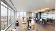 Located in Philadelphia's vibrant North Philadelphia neighborhood, this high-rise amenity lounge at 1717 N 12th St offers a refined and contemporary communal experience. The single-floor space is thoughtfully divided into two living areas, each designed to serve distinct social functions while maintaining a cohesive modern aesthetic throughout.

Floor-to-ceiling curtain-wall glazing wraps around the perimeter, flooding the interior with abundant natural light and framing breathtaking panoramic views of the Philadelphia skyline and surrounding cityscape. The lounge area features plush, neutral-toned seating arrangements — including curved accent chairs, a generous sofa, and low side tables — set against a dramatic full-wall built-in bookcase with warm backlit shelving, creating an inviting library-like atmosphere.

The second living zone transitions into a social and entertainment hub, with high-top bistro tables paired with leather-upholstered barstools in gold and tan tones. A sleek wet bar anchors this area, complete with stone tile backsplash, pendant lighting, and dark cabinetry accented by warm overhead cove lighting. Patterned area rugs and a mix of hard flooring surfaces delineate the zones elegantly. Together, the two areas offer a versatile, upscale environment ideal for both relaxed lounging and social gatherings.