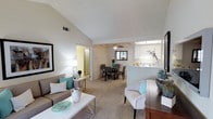 This single-floor residence offers a thoughtfully designed open-concept layout that seamlessly connects the living, dining, and kitchen areas into one cohesive, light-filled space. The living room is anchored by vaulted ceilings that lend a sense of airiness and volume, while plush wall-to-wall carpeting adds warmth underfoot. Neutral tones throughout the space create a calm, versatile backdrop, complemented by tasteful décor accents in teal that provide subtle pops of color.

The living area is generously proportioned, comfortably accommodating a full sofa arrangement and accent seating, and flows naturally into the dining area, which features a ceiling fan and is bathed in natural light from a skylight above. This transition creates an inviting space well-suited for both everyday living and entertaining.

Adjacent to the dining area, the kitchen is positioned behind a half-wall peninsula, offering a functional pass-through design. White cabinetry and clean surfaces give the kitchen a bright, tidy appearance. A window above the sink allows for additional natural light.

A hallway visible from the living area leads to additional rooms, suggesting a practical and well-organized floor plan. Overall, the space delivers a bright, comfortable, and cohesive living environment with a clean, contemporary aesthetic.