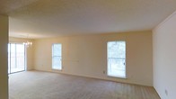 This single-story space features a generously sized main room finished with wall-to-wall carpet in a neutral tone, contributing to a clean and understated aesthetic. The room is defined by its simplicity, with smooth, light-colored walls that create a bright and open atmosphere. Two evenly spaced windows line the far wall, allowing natural light to filter in throughout the day while offering views of surrounding greenery outside. The windows are fitted with horizontal blinds, providing adjustable light control and privacy. A textured ceiling adds a subtle visual element to the otherwise minimalist interior. Electrical outlets are conveniently positioned along the baseboard, indicating practical utility throughout the space. The neutral palette of the carpet, walls, and ceiling work cohesively to create a versatile backdrop that can accommodate a variety of furnishing styles. The overall ambiance is calm and understated, well-suited for comfortable everyday living.