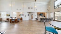 Located at 2400 N Morrison Blvd in Hammond, Louisiana, this single-floor clubhouse offers a generously proportioned and thoughtfully designed communal space. The centerpiece of the facility is a dramatic open-plan living area defined by soaring vaulted ceilings with exposed dark wood truss detailing and an abundance of natural light streaming in through large clerestory windows and full-height glass panels flanking the entryway. Warm wood-look flooring runs throughout the main area, complementing the bright white walls and contemporary furnishings. A standout stacked-stone accent wall anchors the lounge area, where a wall-mounted TV, granite-topped bar counter with stone base, and built-in cabinetry create an inviting gathering space. A living green wall adds a striking natural element to the interior. The reception area near the entrance features comfortable seating, artwork, and a wood-toned front desk. A dedicated office room offers a computer workstation lineup along a window wall with views of surrounding greenery, finished with patterned carpet tile flooring. An additional flexible room with a whiteboard, sofa, and interior window overlooking the main hall serves well as a meeting or study space. Glass doors open to an outdoor area, further enhancing the sense of openness and connectivity throughout this well-appointed facility.