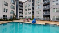 Located at 1608 E Riverside Dr in Austin, Texas, this outdoor amenity space is designed to deliver a resort-style living experience within a contemporary multi-story residential community. The centerpiece is a spacious, sparkling swimming pool with a wide surrounding deck outfitted with lounge chairs, perfect for relaxing in the Texas sun. The pool area is enclosed by the community's modern building facades, featuring large windows, private balconies, and a clean mix of gray and white exterior finishes that lend the space a sleek, urban aesthetic.

Adjacent to the pool, a covered pergola provides a shaded retreat anchored by a well-appointed outdoor kitchen. The kitchen features dark granite countertops set atop a classic brick base, stainless steel grills, a built-in sink, and additional prep surfaces — ideal for outdoor entertaining. The pergola's overhead structure frames open views of the surrounding buildings and sky, creating a seamless blend of shelter and open-air ambiance.

Brick-paved flooring throughout the outdoor kitchen and lounge areas adds warmth and texture to the space. Lush landscaping with young trees and plantings borders the pool deck, softening the hardscape and enhancing the overall outdoor environment. A mounted TV near the pool adds a recreational touch, rounding out this comprehensive amenity area.