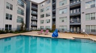 Located at 1608 E Riverside Dr in Austin, Texas, this outdoor amenity space is designed to deliver a resort-style living experience within a contemporary multi-story residential community. The centerpiece is a spacious, sparkling swimming pool with a wide surrounding deck outfitted with lounge chairs, perfect for relaxing in the Texas sun. The pool area is enclosed by the community's modern building facades, featuring large windows, private balconies, and a clean mix of gray and white exterior finishes that lend the space a sleek, urban aesthetic.

Adjacent to the pool, a covered pergola provides a shaded retreat anchored by a well-appointed outdoor kitchen. The kitchen features dark granite countertops set atop a classic brick base, stainless steel grills, a built-in sink, and additional prep surfaces — ideal for outdoor entertaining. The pergola's overhead structure frames open views of the surrounding buildings and sky, creating a seamless blend of shelter and open-air ambiance.

Brick-paved flooring throughout the outdoor kitchen and lounge areas adds warmth and texture to the space. Lush landscaping with young trees and plantings borders the pool deck, softening the hardscape and enhancing the overall outdoor environment. A mounted TV near the pool adds a recreational touch, rounding out this comprehensive amenity area.