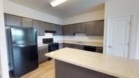 This single-floor space showcases a well-appointed kitchen designed with both functionality and modern aesthetics in mind. The kitchen features an abundance of dark espresso-toned cabinetry that extends from floor to ceiling, providing generous storage throughout. The cabinetry is complemented by sleek brushed metal hardware, adding a refined touch to the overall design.

Light-toned countertops offer a striking contrast against the dark cabinets, creating a balanced and visually appealing palette. A spacious kitchen island anchors the space, providing additional prep area and serving as a natural focal point. The flooring consists of warm wood-tone laminate planks, adding a welcoming warmth underfoot.

The kitchen is fully equipped with a suite of black appliances, including a refrigerator, electric range with oven, and dishwasher, all seamlessly integrated into the cabinetry layout. An under-cabinet range hood is positioned above the cooktop for practical ventilation. The sink is centrally located along the counter run, surrounded by ample workspace on either side.

Overhead fluorescent lighting ensures the kitchen is well-lit, enhancing the clean, bright feel of the space. The open layout and thoughtful design make this kitchen both practical for everyday use and inviting in appearance.